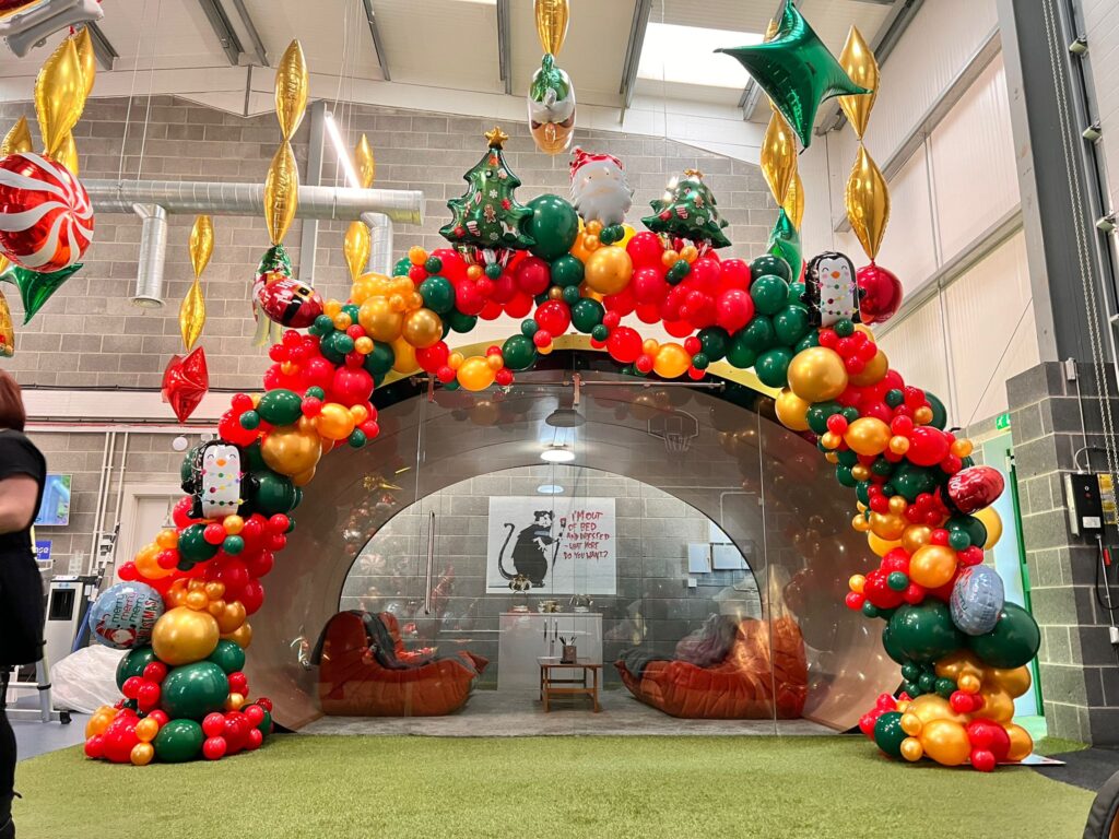 Large organic Christmas balloon arch in red, green, and gold framing a glass office pod, decorated with festive foil balloons including trees, stars, penguins, and Santa, installed in a modern industrial workspace.